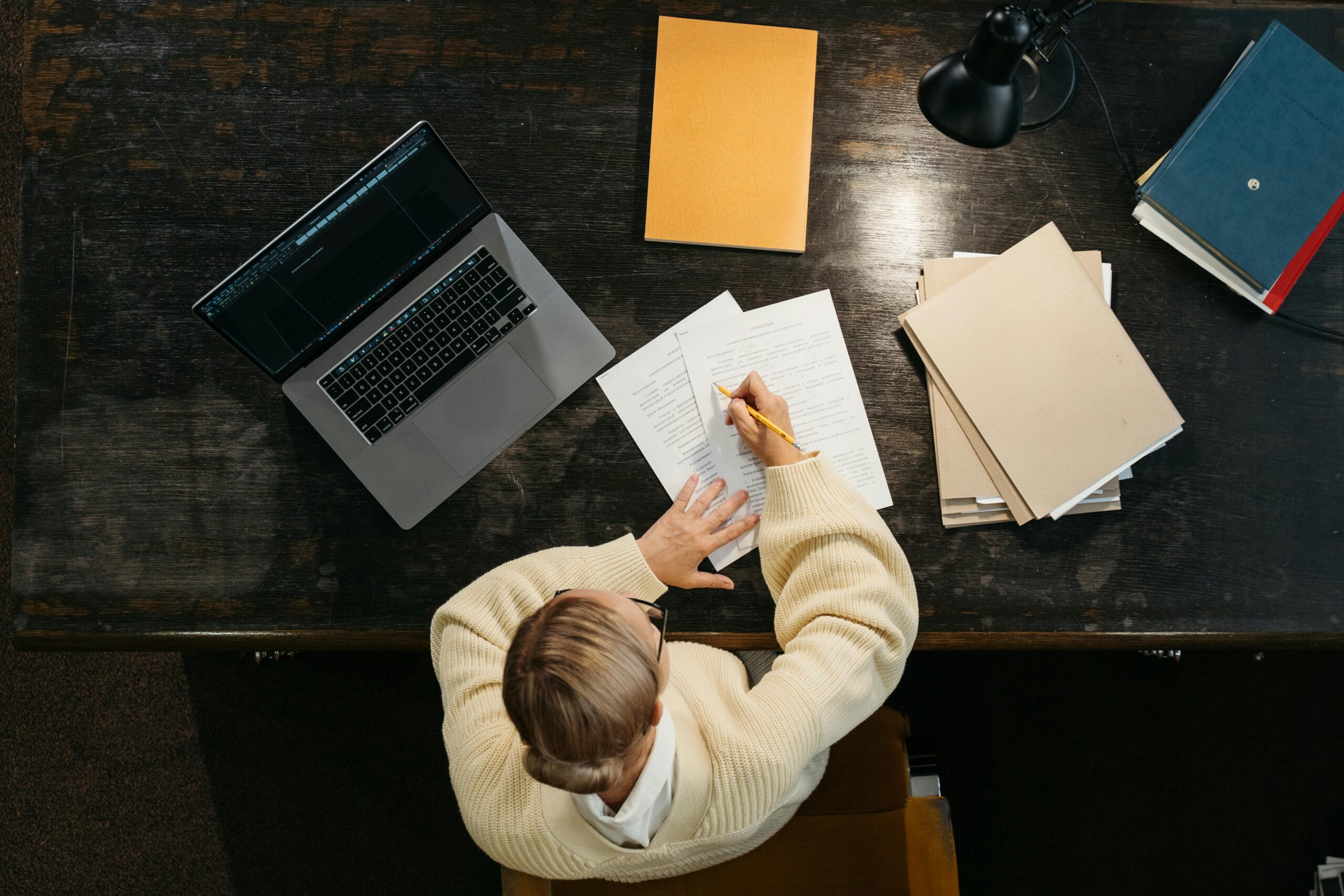 Overhead view of woman writing at desk with laptop, papers, and books.